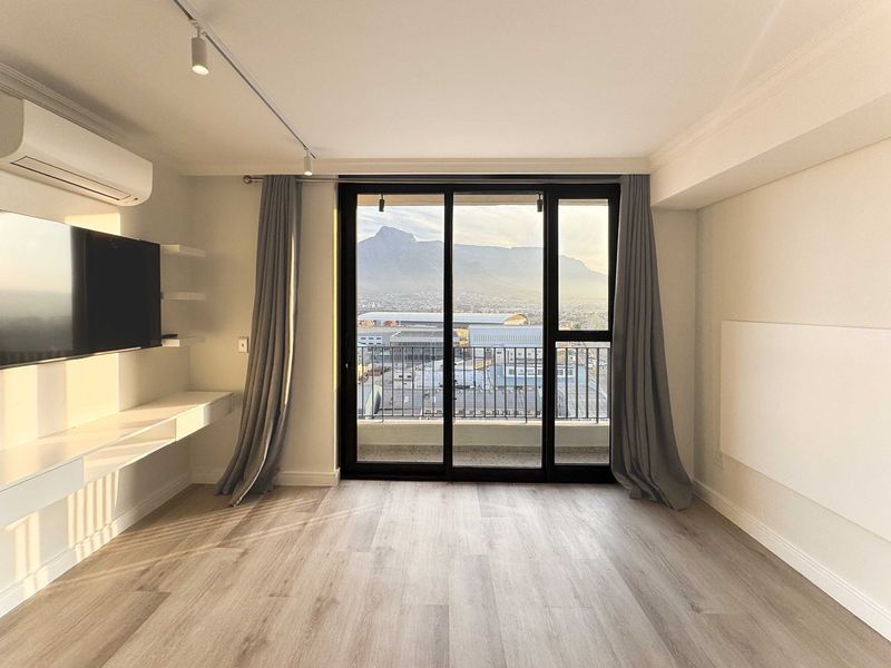 Bright and modern living area featuring light laminate flooring, track lighting, built-in shelving, and a sliding glass door leading to a balcony with mountain views.