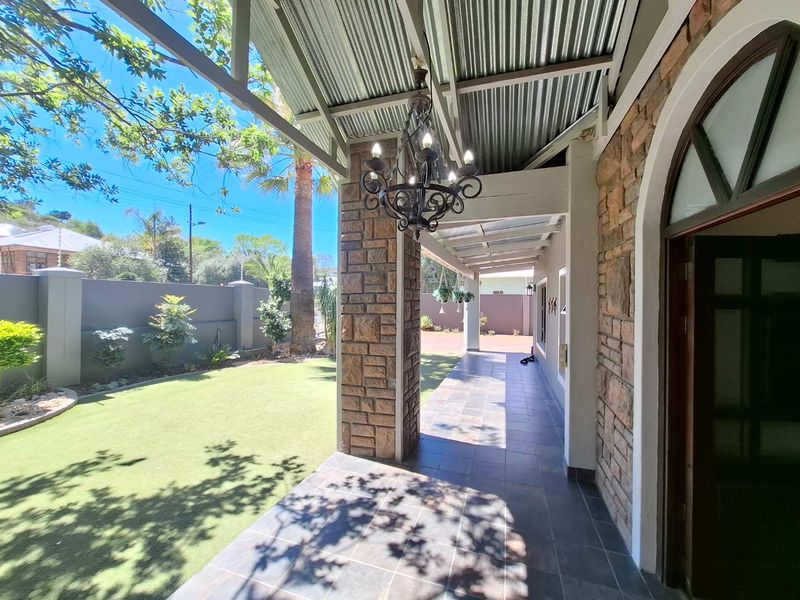 A covered patio area featuring stone-clad pillars, tiled flooring, and a decorative chandelier, overlooking a well-maintained garden with a boundary wall.
