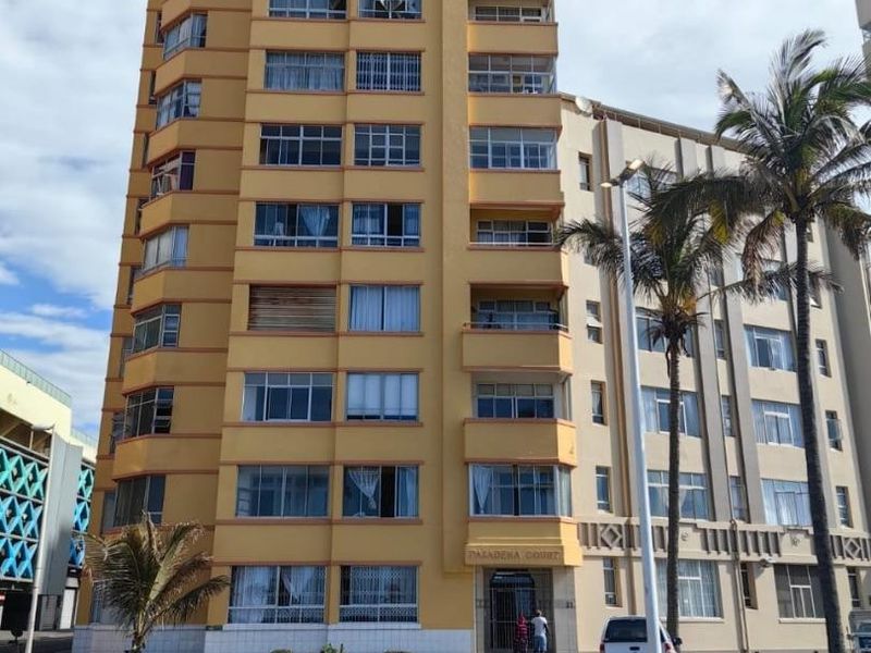 A multi-story apartment building facade featuring balconies and a ground-level entrance, situated on a corner lot with street access.