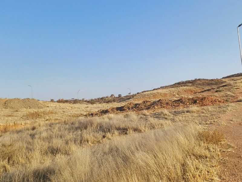 A view of a vacant residential plot featuring natural dry grass terrain, adjacent to a paved road with existing street lighting infrastructure.