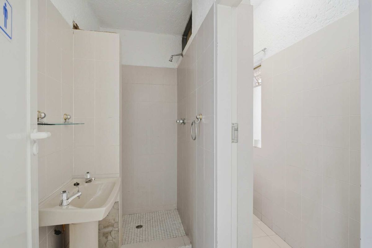 A functional office bathroom featuring a pedestal sink, tiled shower enclosure, and neutral wall tiling, requiring minor cosmetic updates.
