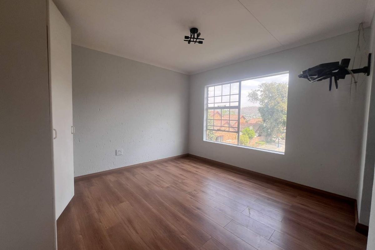 A bright, empty bedroom featuring wood-look laminate flooring, built-in cupboards, and a large window with neighbourhood views.
