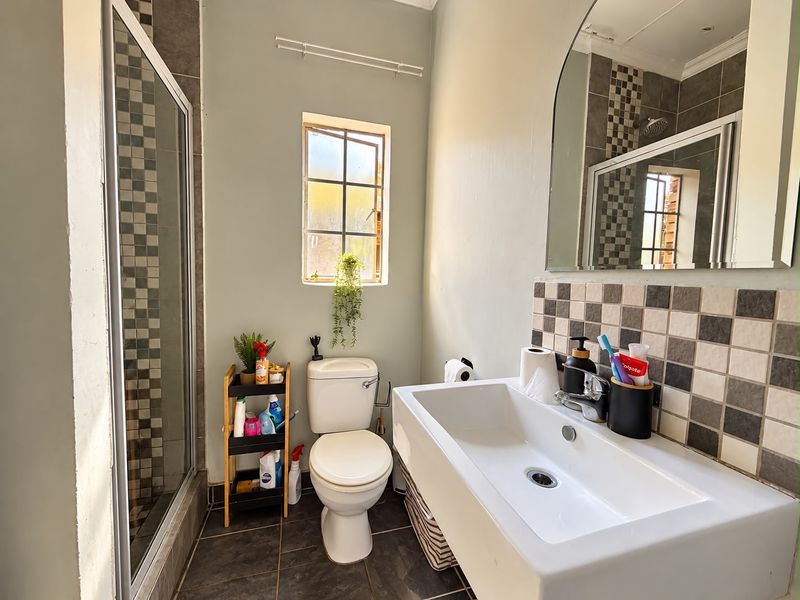 A functional bathroom featuring a shower enclosure, wall-mounted basin with tiled splashback, and a toilet, finished with neutral wall paint and tiled flooring.