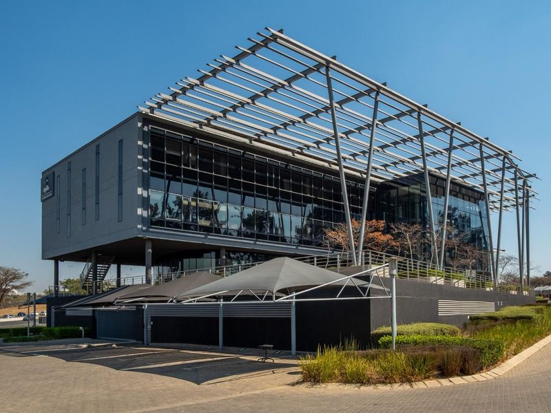 Modern multi-story office building featuring a prominent steel pergola structure, extensive glass facade, and paved parking area with shade sails.