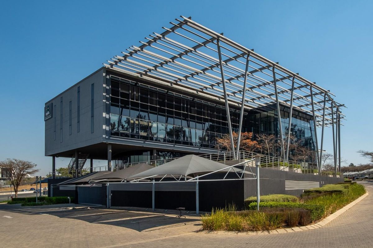 Modern multi-story office building featuring a prominent steel pergola structure, extensive glass facade, and paved parking area with shade sails.