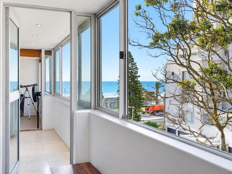 A bright, enclosed balcony featuring tiled flooring, recessed lighting, and expansive windows offering clear ocean views.