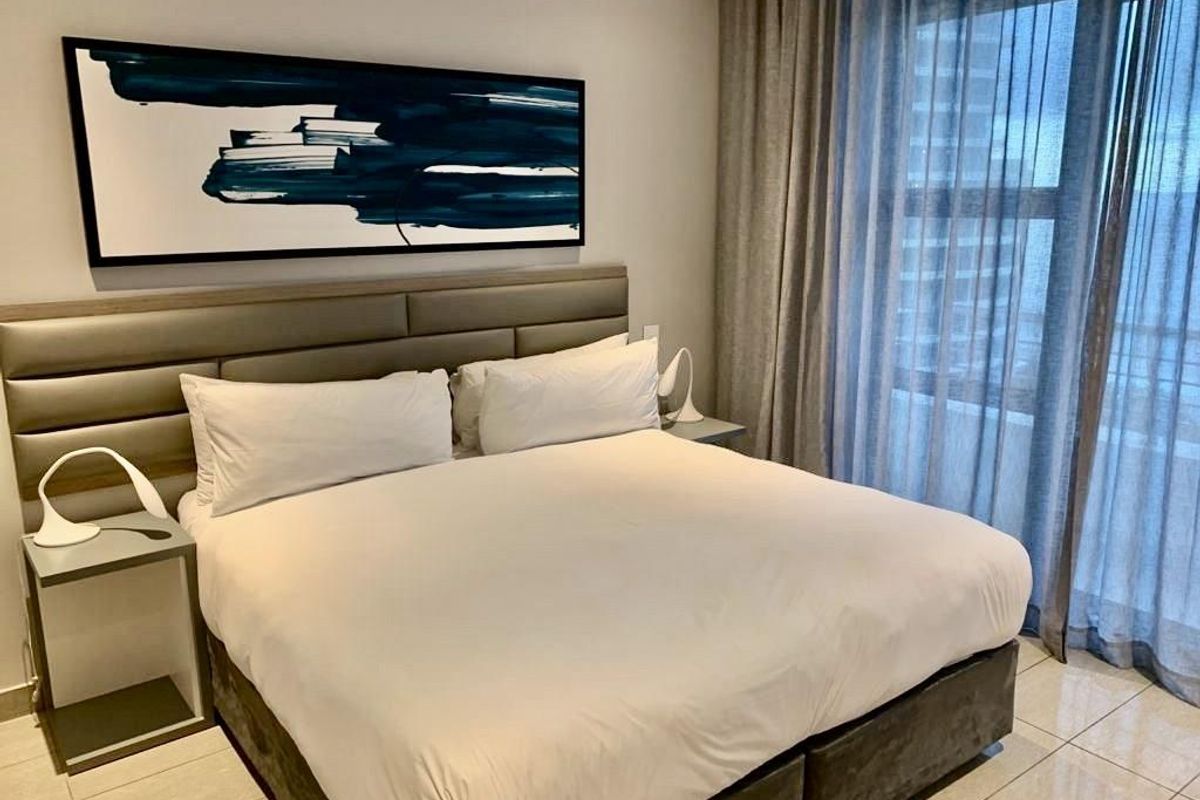 A modern bedroom featuring a built-in upholstered headboard, tiled flooring, and floor-to-ceiling curtains leading to a balcony.