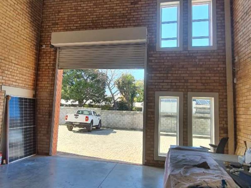 Spacious industrial utility area featuring high-volume ceilings, exposed brickwork, concrete flooring, and a large roller shutter door for easy access.