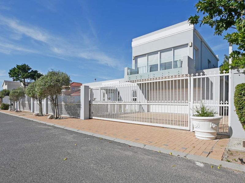 Front exterior view of a modern apartment building featuring a secure metal gate, paved driveway, and an upper-level balcony with glass balustrades.