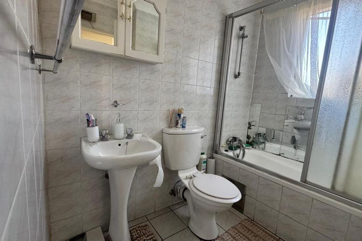 A functional bathroom featuring a pedestal basin, toilet, and a combined bathtub with shower screen, finished with neutral wall and floor tiling.