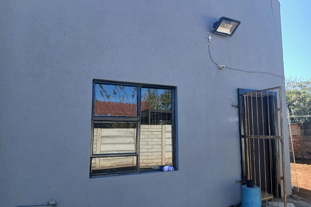 Exterior view of the cottage rear facade featuring a security gate, metal-framed window, and mounted floodlight on a painted masonry wall.