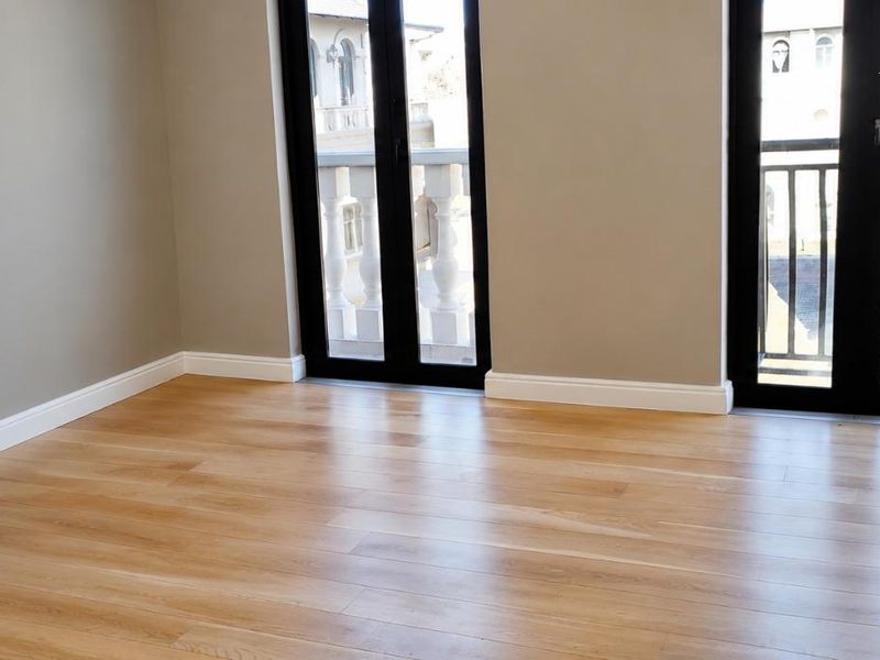 A bright, empty bedroom featuring light wood laminate flooring, neutral walls, and two sets of double doors opening onto a balcony.