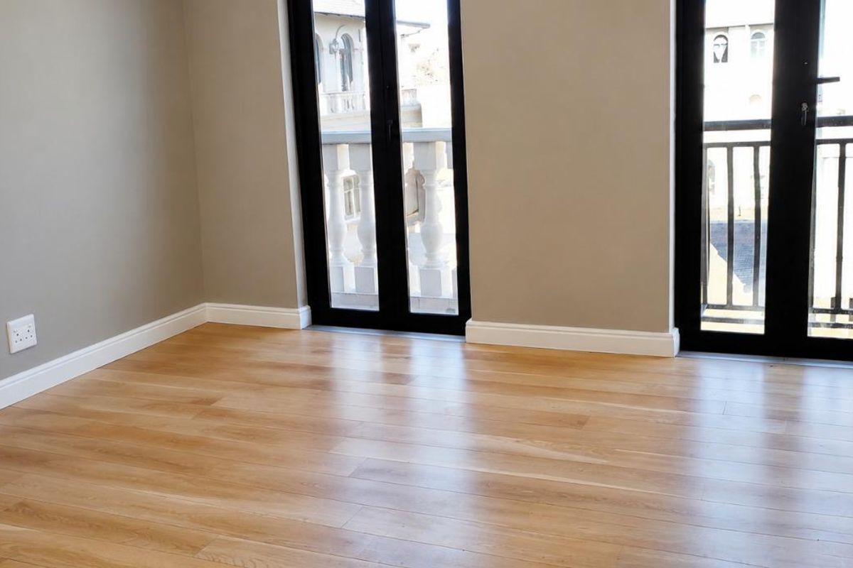 A bright, empty bedroom featuring light wood laminate flooring, neutral walls, and two sets of double doors opening onto a balcony.