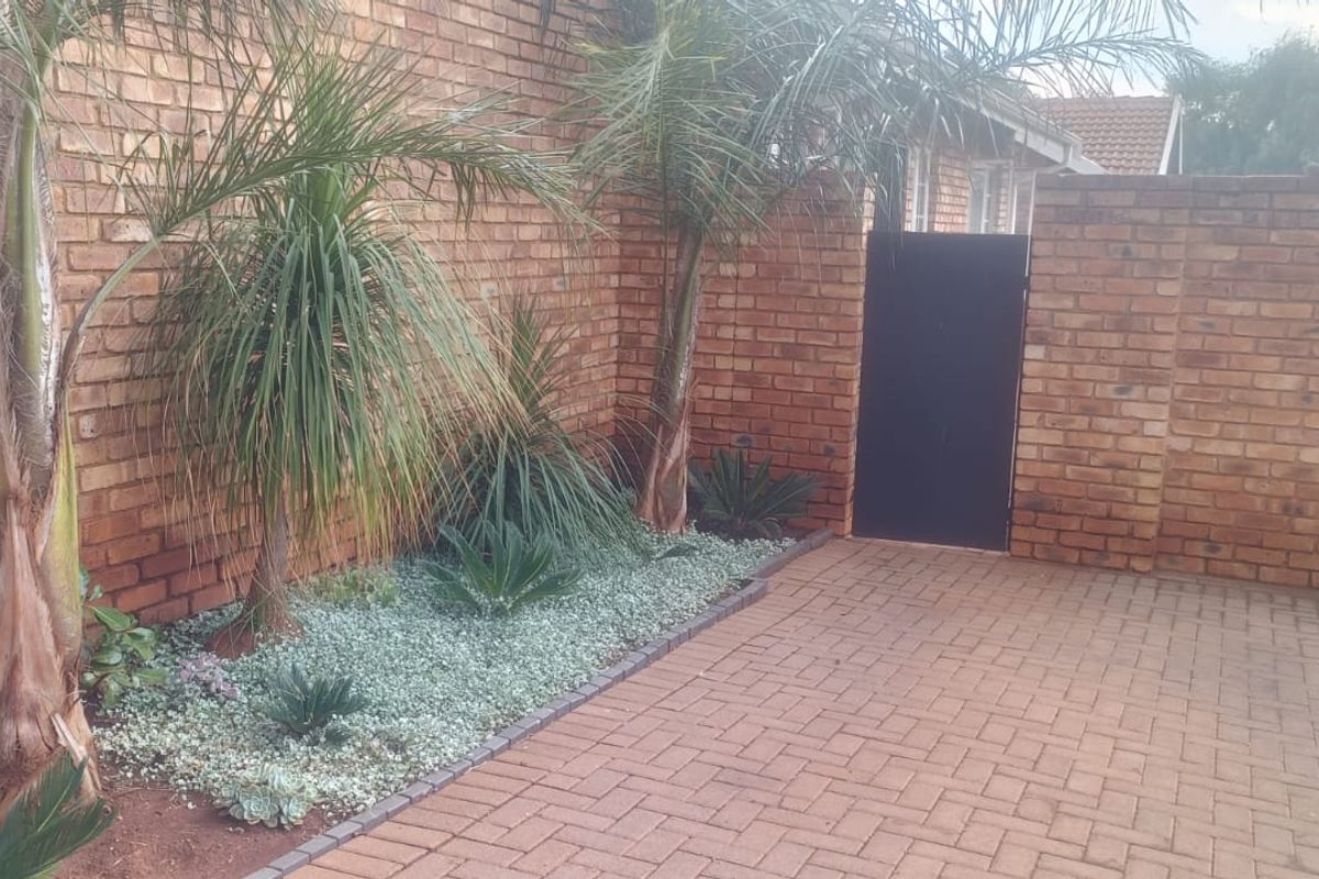 A private courtyard area featuring brick paving, a secure metal gate, and established garden beds with palm trees and low-maintenance ground cover.
