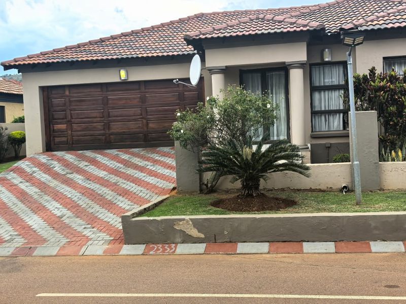 Front elevation of a single-story house featuring a paved driveway, wooden garage door, and a low boundary wall with established garden.