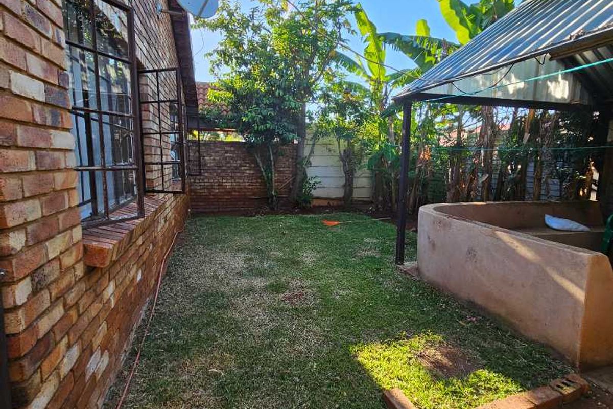 A rear garden area featuring a brick exterior wall, a small lawn, and a covered outdoor structure with a built-in masonry feature.