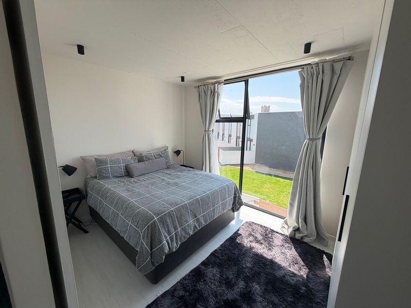 A bright bedroom featuring an exposed concrete ceiling, recessed spotlights, and a large floor-to-ceiling window with views of the surrounding area.