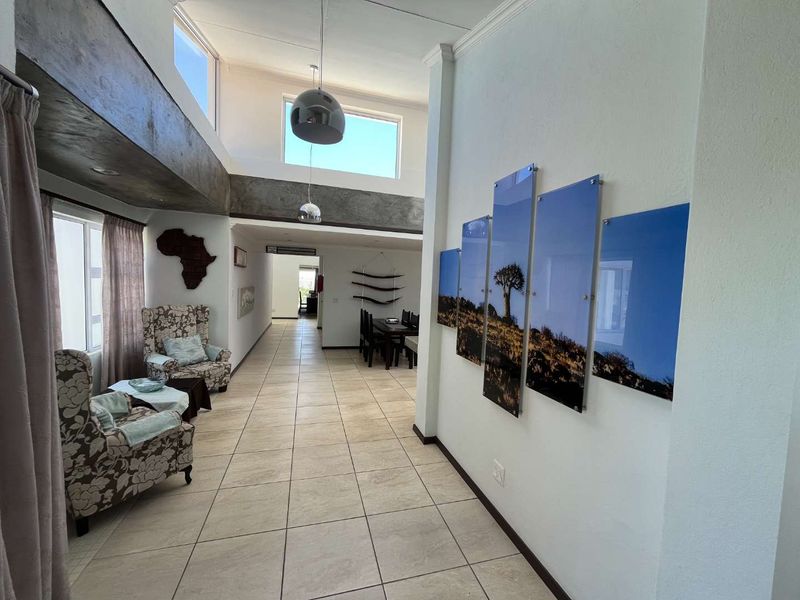 Spacious open-plan living area featuring high double-volume ceilings with exposed concrete beams, large windows for natural light, and neutral tiled flooring throughout.