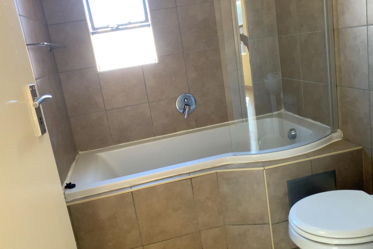 A functional bathroom featuring a tiled bathtub with a glass shower screen, a wall-mounted mixer, and a standard toilet.