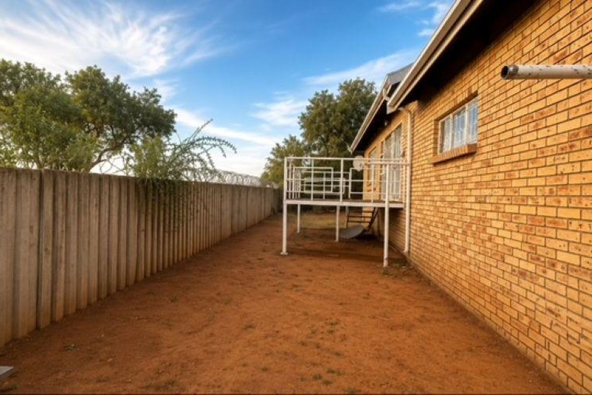 Rear exterior view of the property featuring a brick facade, a raised metal balcony, and a large, unlandscaped backyard area with boundary walling.