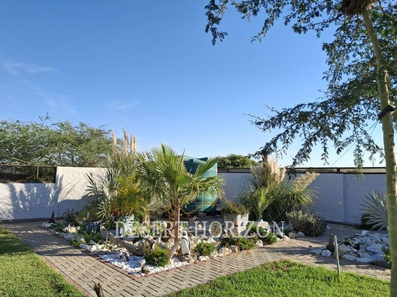 A well-maintained garden area featuring a paved walkway, landscaped beds with decorative stones, and a large water storage tank against a boundary wall.