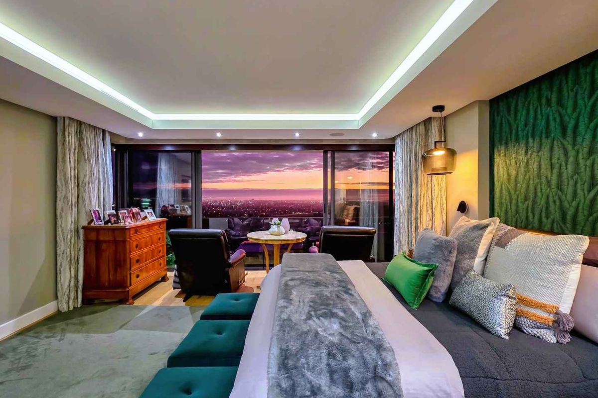 A luxurious bedroom featuring recessed tray ceiling lighting, floor-to-ceiling sliding glass doors, and a private balcony with expansive sunset city views.