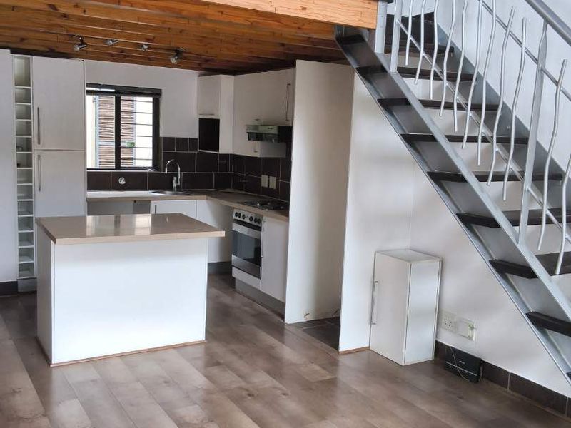Open-plan living area featuring a modern kitchen with island, laminate flooring, and a staircase leading to a mezzanine level with exposed timber beams.