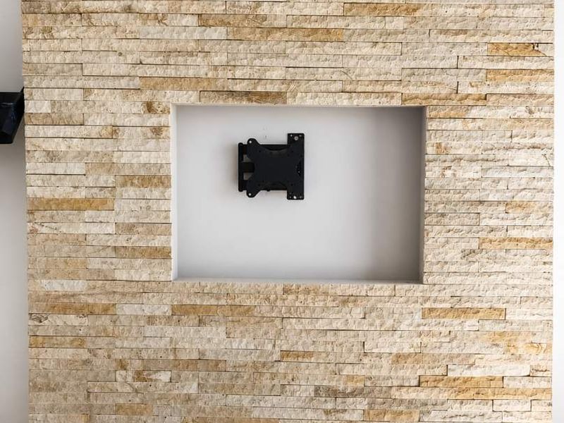A feature wall finished in natural stacked stone, featuring a recessed niche with a mounted television bracket, set above tiled flooring.