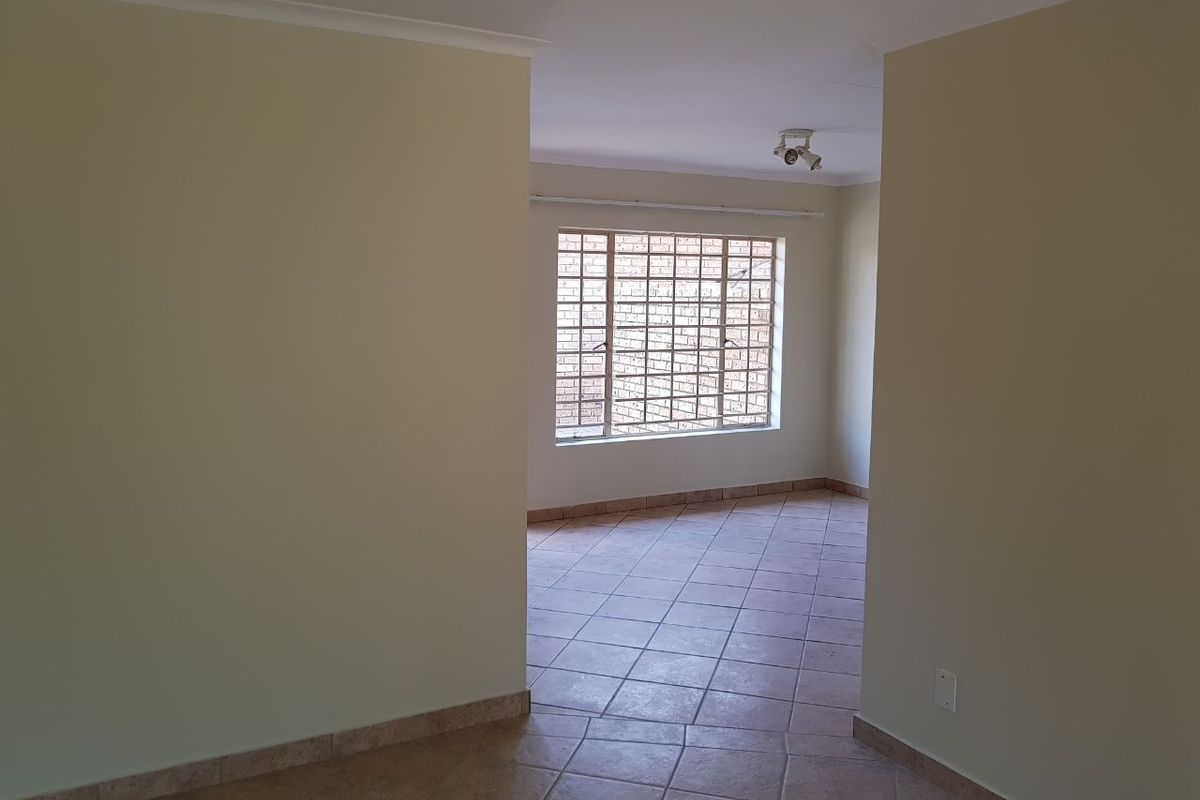 An empty, open-plan living area featuring neutral tiled flooring, a large window with security bars, and simple ceiling lighting.