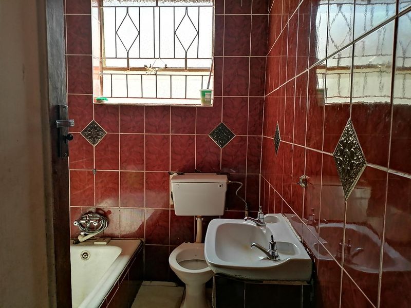A compact bathroom featuring dated red wall tiles, a standard bathtub, pedestal basin, and toilet, with security bars on the window.