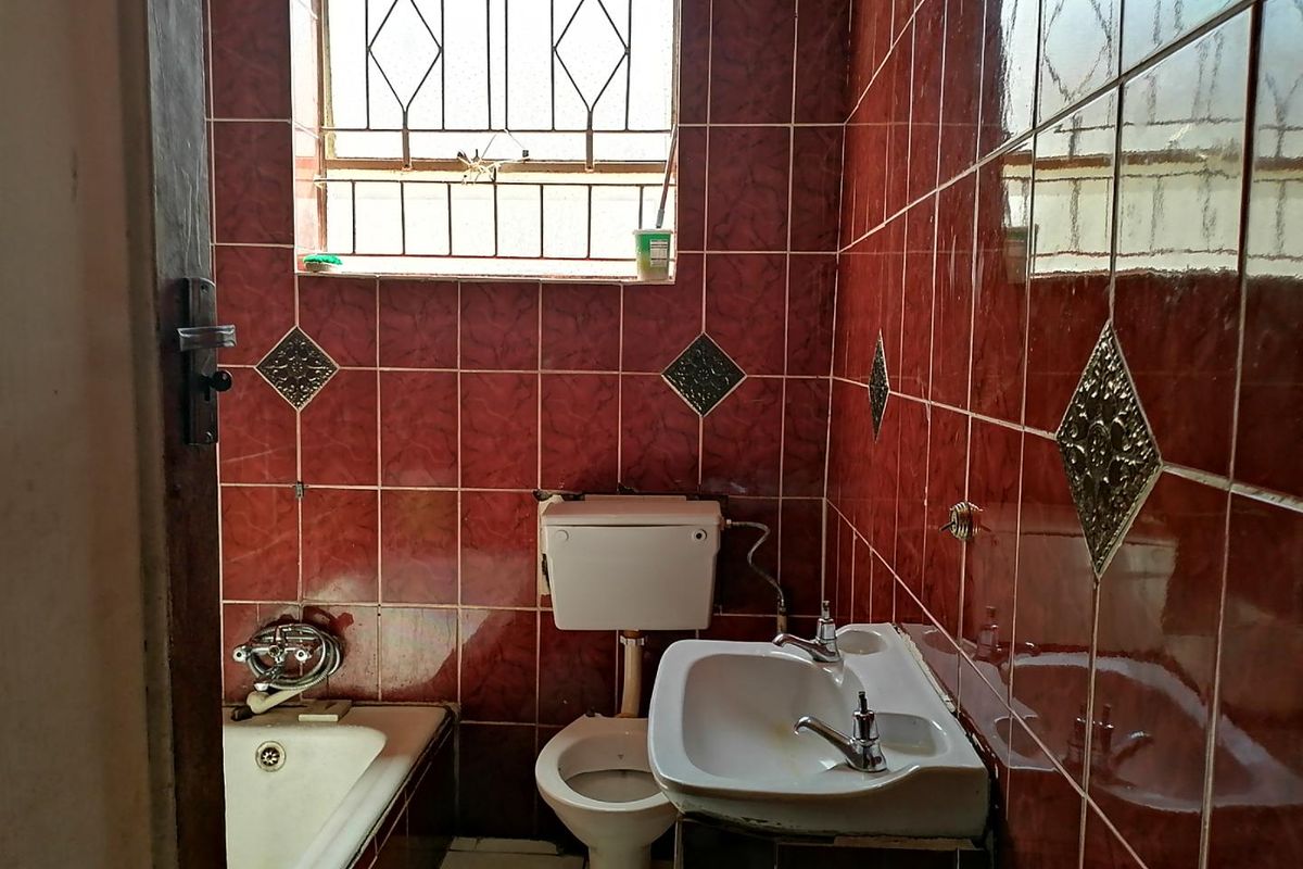 A compact bathroom featuring dated red wall tiles, a standard bathtub, pedestal basin, and toilet, with security bars on the window.