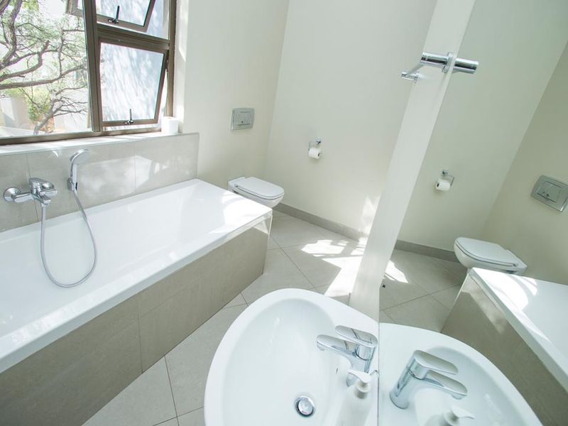 Modern bathroom featuring a built-in bathtub, wall-mounted toilet, vanity basin with chrome fixtures, and large mirror, finished with neutral floor tiles.