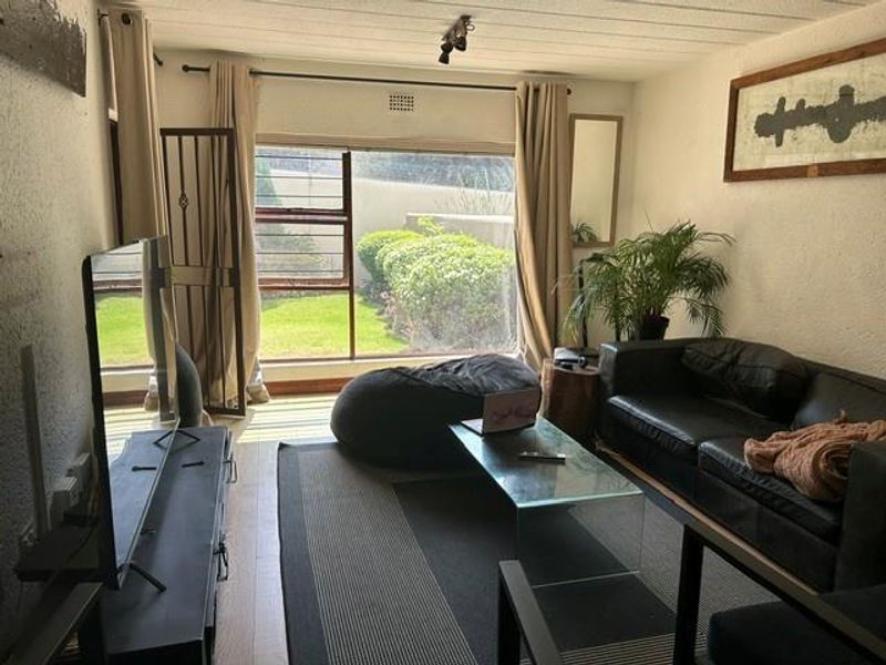 A living room featuring large windows with garden views, textured walls, and laminate flooring, providing a functional space with natural light.