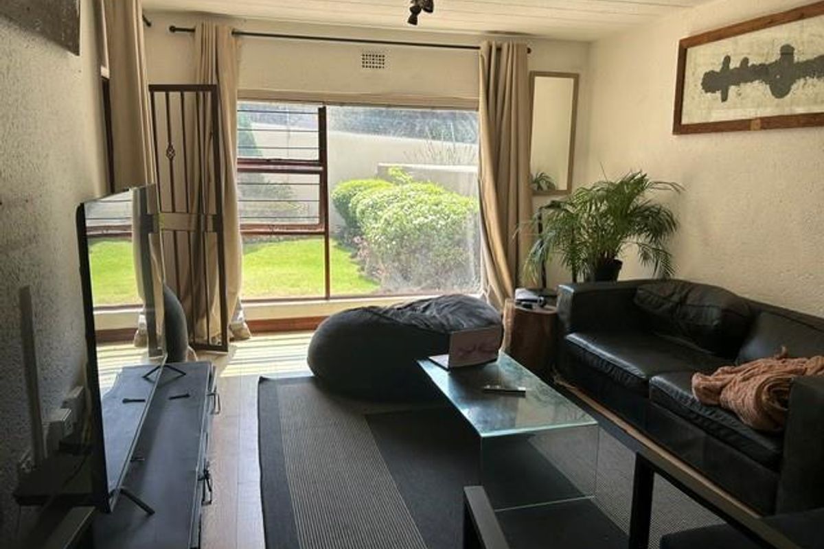 A living room featuring large windows with garden views, textured walls, and laminate flooring, providing a functional space with natural light.