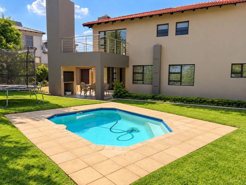 Rear view of the property featuring a swimming pool, paved surround, covered patio with built-in braai, and an upper-level balcony.