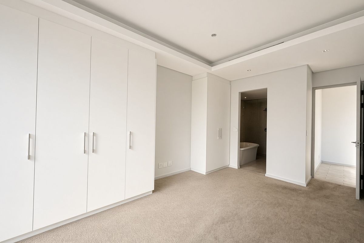 Spacious bedroom featuring extensive built-in cupboards, neutral carpeting, and a recessed ceiling with downlights, leading into an en-suite bathroom.