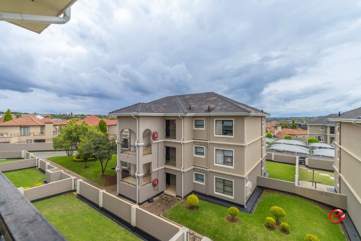An elevated aerial view of a multi-story apartment building featuring a tiled roof, beige facade, and surrounding landscaped garden areas with paved walkways.