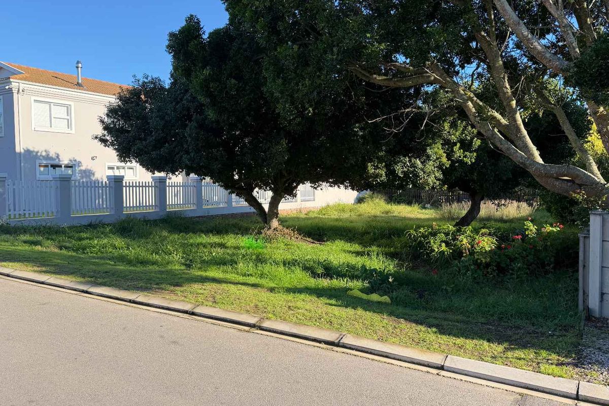 A vacant residential plot featuring mature trees and a white picket fence, situated along a paved street.
