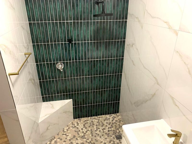 Modern bathroom shower area featuring a striking green tiled feature wall, marble-look wall tiles, pebble flooring, and elegant gold fixtures.