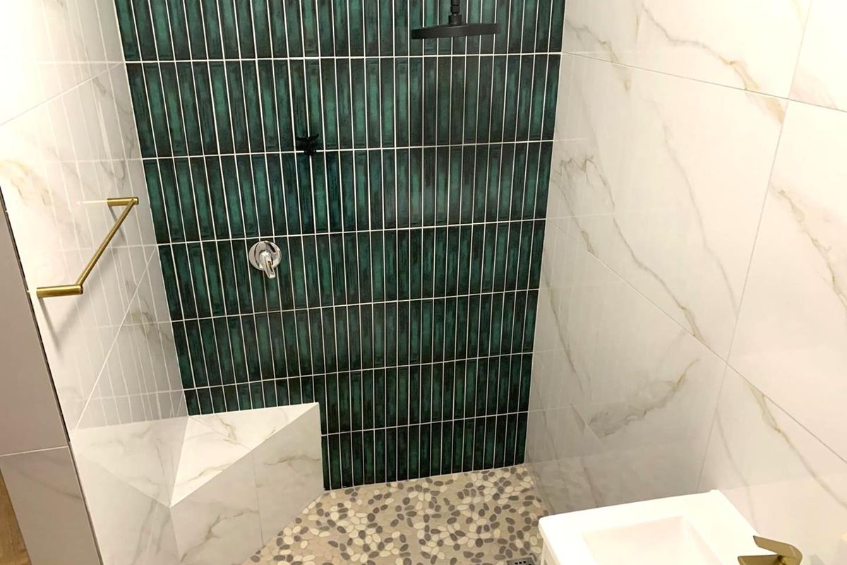 Modern bathroom shower area featuring a striking green tiled feature wall, marble-look wall tiles, pebble flooring, and elegant gold fixtures.