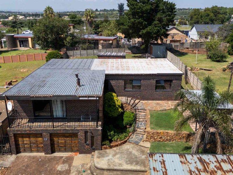 Aerial view of a brick house featuring a double garage, balcony, stone retaining walls, and a large surrounding garden area.