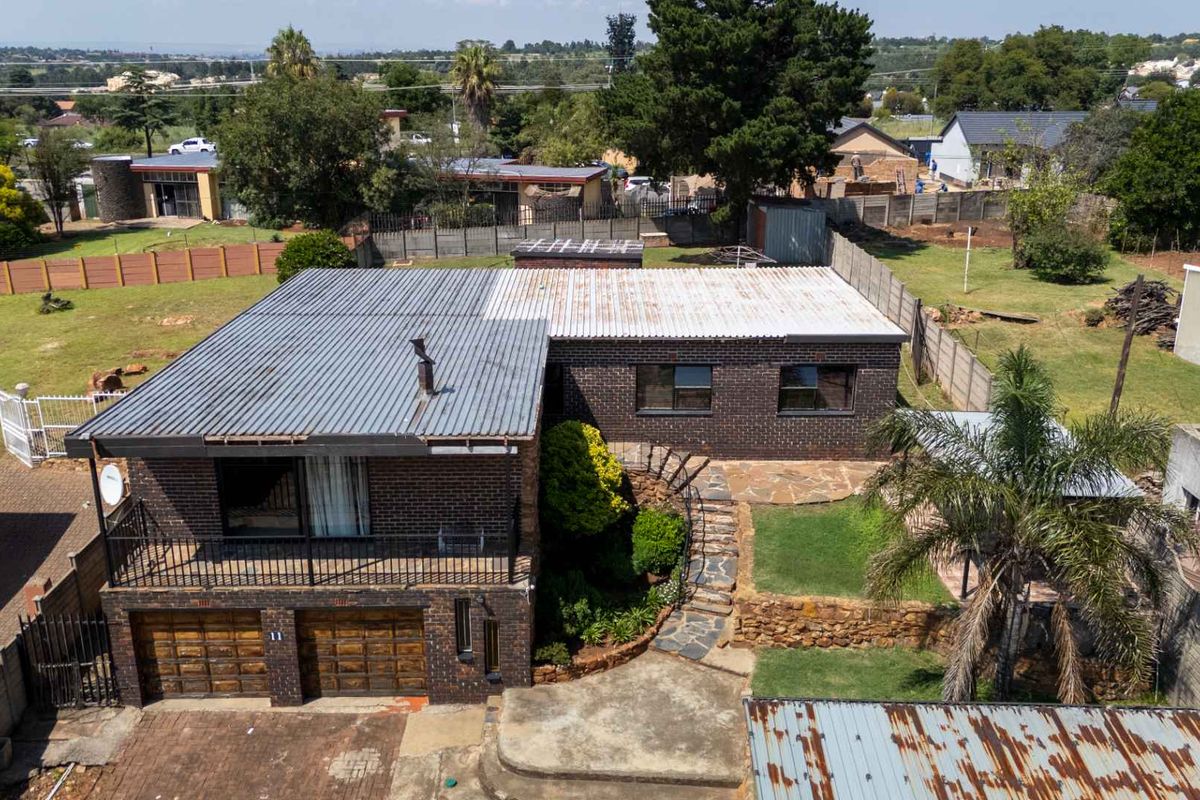 Aerial view of a brick house featuring a double garage, balcony, stone retaining walls, and a large surrounding garden area.