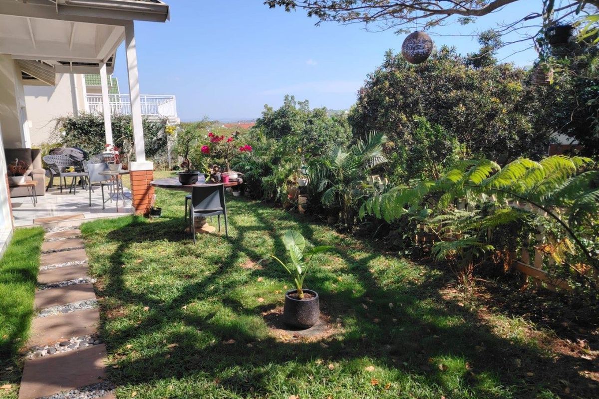 A well-maintained garden area featuring a covered patio, stone paver pathway, and established landscaping with mature trees and shrubs.