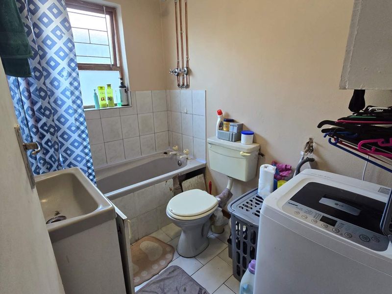 A functional bathroom featuring a bathtub, toilet, and tiled splashback, with exposed plumbing and dated finishes throughout.