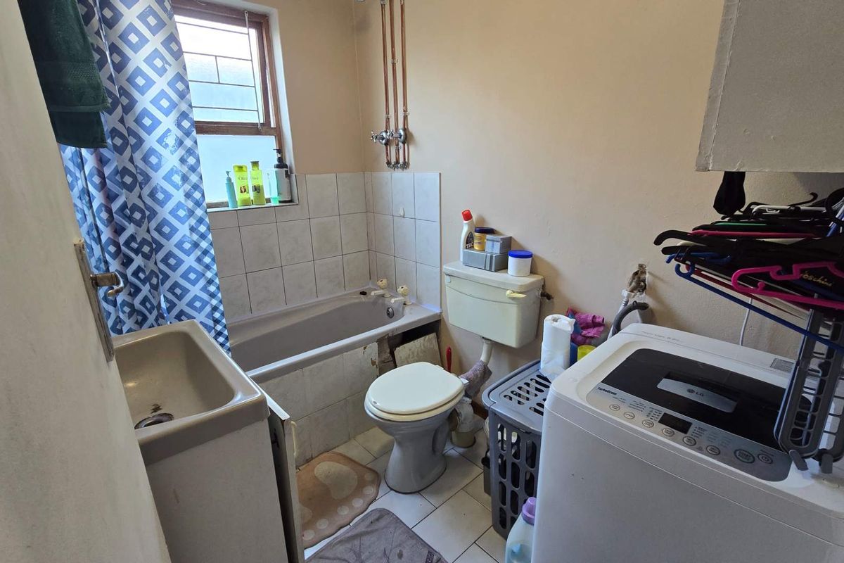 A functional bathroom featuring a bathtub, toilet, and tiled splashback, with exposed plumbing and dated finishes throughout.