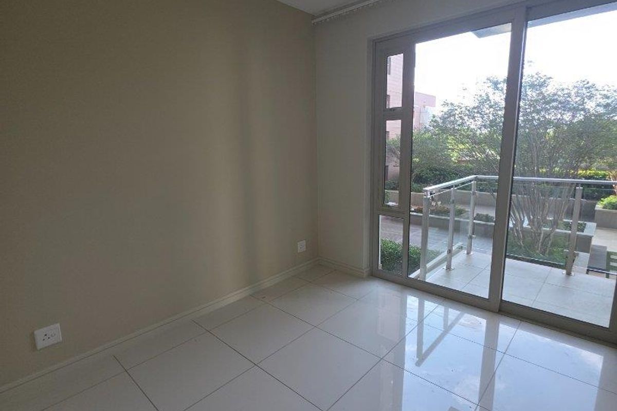 A bright, empty bedroom featuring large sliding glass doors that open onto a private balcony, finished with neutral tiled flooring.