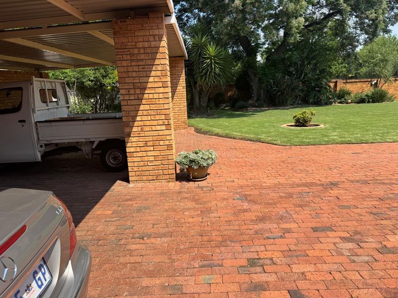 A covered carport area featuring sturdy brick pillars and a paved driveway, adjacent to a well-maintained garden space.