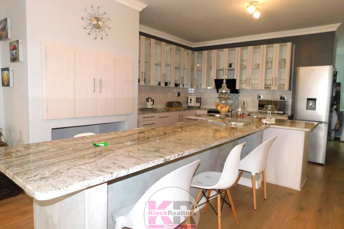 Modern kitchen featuring a large granite-topped breakfast island, ample cabinetry with glass-fronted upper units, and integrated lighting.