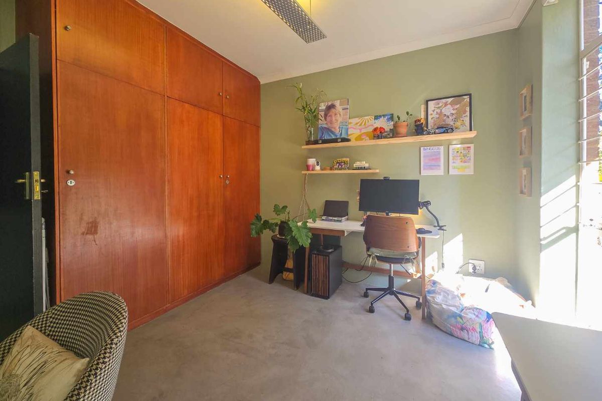 A bedroom featuring floor-to-ceiling built-in wooden cupboards, louvered windows, and neutral flooring, currently utilized as a home office space.