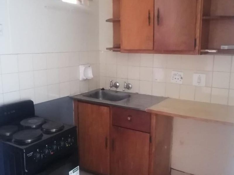 A compact kitchen featuring dated wooden cabinetry, a stainless steel sink, tiled splashback, and a freestanding electric stove.